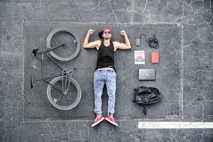 A man lying on a lot, with his bike on one side laid beside him and his bag, phone, laptop and other accessories laid on the other side