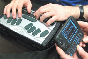 Two people communicating using assistive technology for individuals who are deafblind. One person types a message on a keyboard, while the other person reads it by placing their hand on the device's refreshable braille display