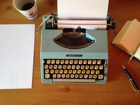 Wooden desk setup featuring a blue typewriter alongside a coffee cup, pen, and notebook