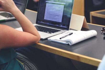 A person at a desk using a laptop, with an open notebook and a pen placed beside them on the table